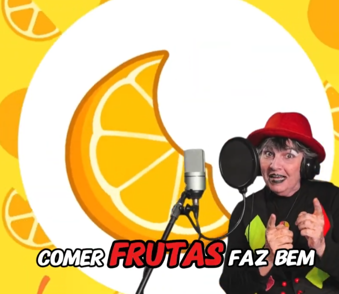 Imagem de divulgação do clipe musical Comer Bem Faz Bem ("Comer Frutas Faz Bem"), com uma artista de chapéu vermelho cantando em frente a um microfone e uma ilustração de laranja ao fundo.