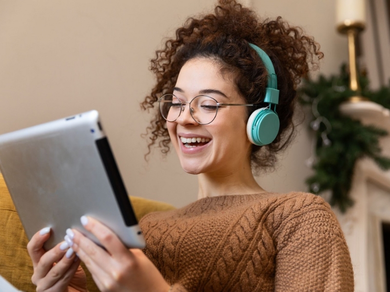 Audiodescritor - Uma jovem com cabelo encaracolado e óculos, vestindo um suéter marrom, sentada e sorrindo. Ela usa fones de ouvido azuis e segura um tablet prateado na mão.