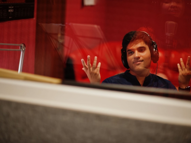 Audiodescrição em Eventos - 
Descrição da Imagem para Texto Alternativo (Alt Text)
Esta imagem é uma fotografia de um homem trabalhando dentro de uma cabine de estúdio, visto através de uma janela.