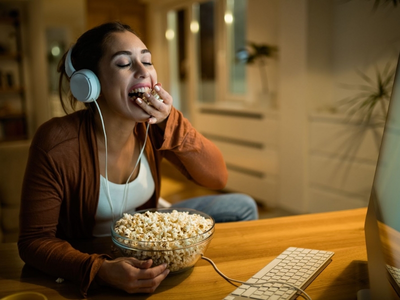 Audiodescrição em Filmes - Esta imagem é uma fotografia que captura uma mulher desfrutando de um momento de relaxamento enquanto assiste a algo em uma tela e come pipoca.