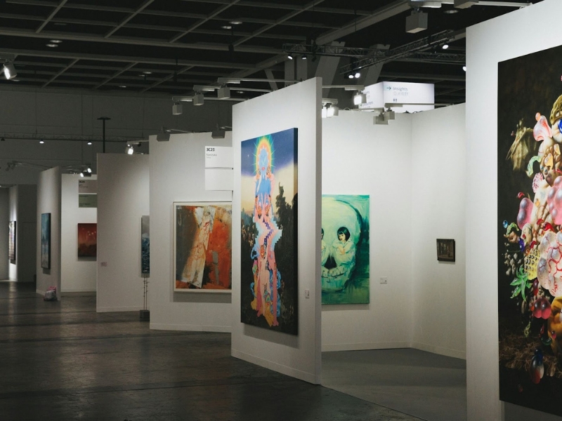 Interior moderno de uma feira ou galeria de arte com paredes brancas em cubículos, exibindo várias pinturas coloridas de arte contemporânea. Audiodescrição em Exposições de Arte