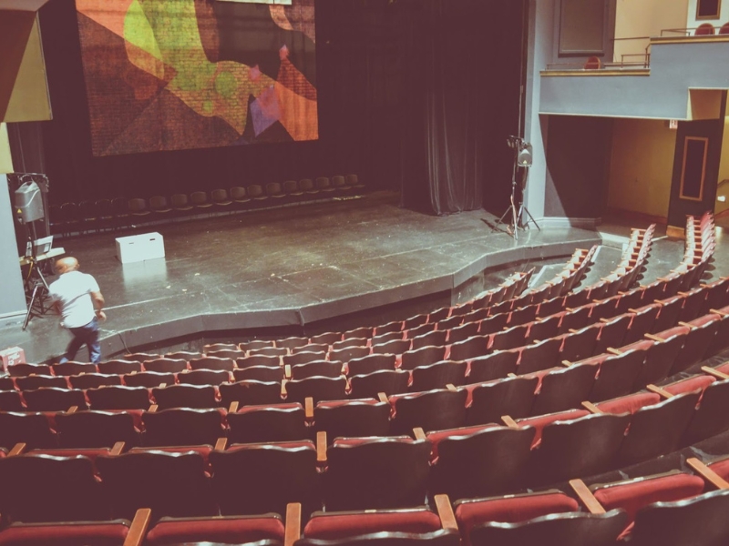 Vista de cima de um teatro vazio com poltronas vermelhas curvas e um homem no palco escuro com projeção abstrata colorida ao fundo.

Importância da Audiodescrição no Teatro