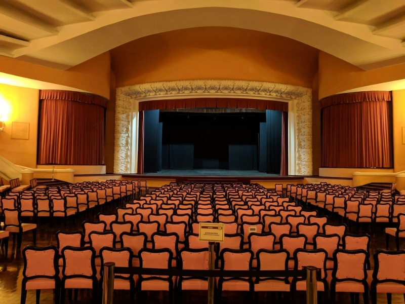 Interior de um teatro clássico vazio com fileiras de poltronas rosas e pretas e um palco com cortinas de veludo vermelho e arco ornamentado. Importância da Audiodescrição no Teatro