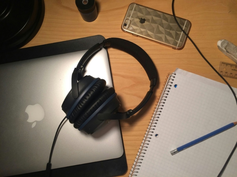 Fones de ouvido pretos sobre um MacBook Pro prateado, com um iPhone dourado e caderno em uma mesa de madeira clara. Qual a diferença entre audiodescrição e autodescrição?